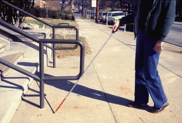 A visually impaired person with a cane walks on a pavement